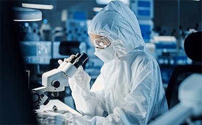 A scientist wearing full PPE observes a sample on a microscope.