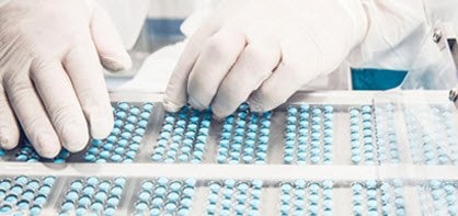 A pharmaceutical or medical manufacturing facility. It shows a person in protective clothing handling trays filled with blue capsules.