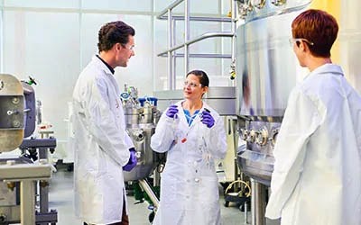 Three individuals in lab coats are in a well-equipped laboratory setting. The person in the center is engaged in a conversation with the individual on the left. The person on the right is observing.