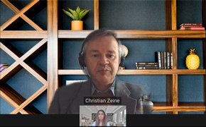 A virtual meeting screenshot with Christian Zane and another participant. A small window shows the other participant speaking. In the background, a well-organized shelf holds books, decorative objects, and a potted plant.