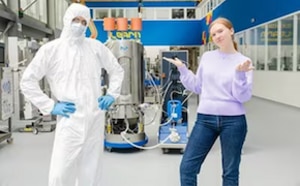 Two scientists, standing in a lab, a male in bunny suit, a female wearing casual clothes